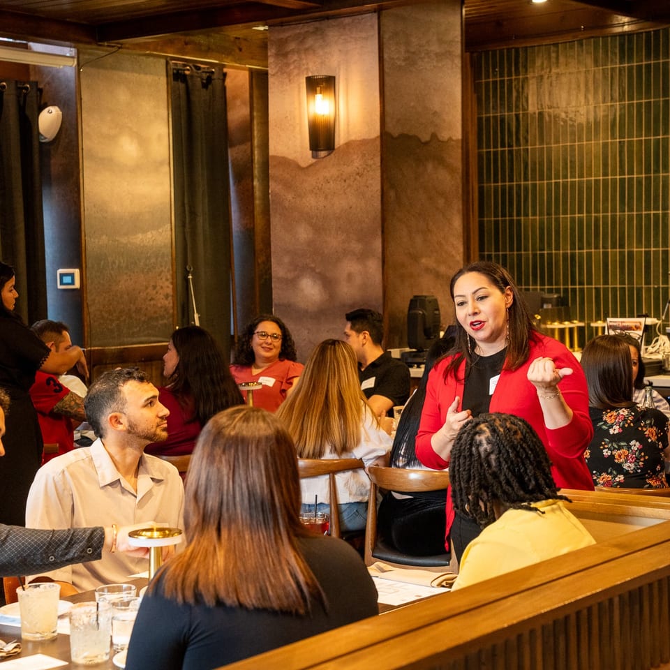 Woman in red jacket speaks to guests at tables in a cozy restaurant with warm lighting.