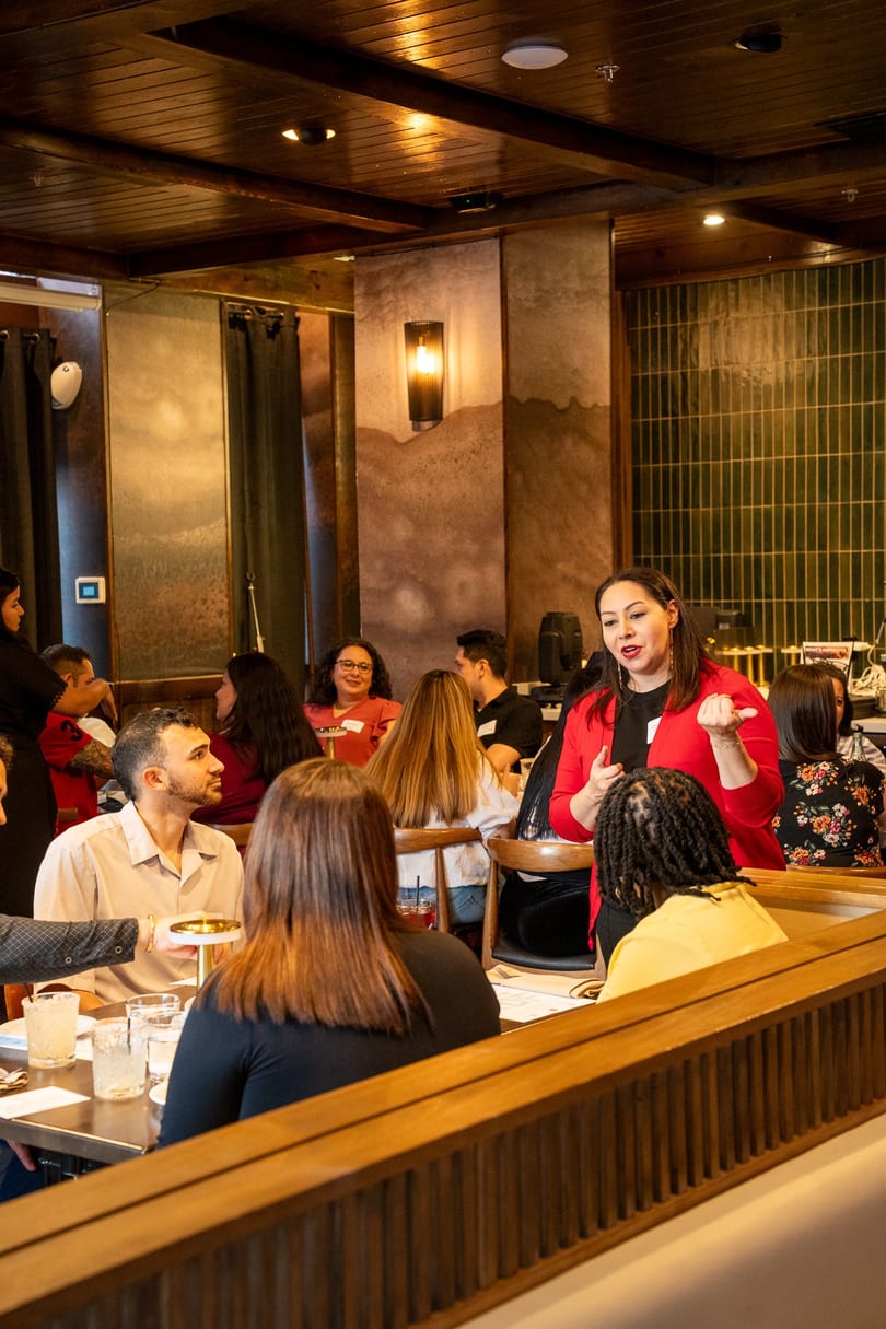 Woman in red jacket speaks to guests at tables in a cozy restaurant with warm lighting.
