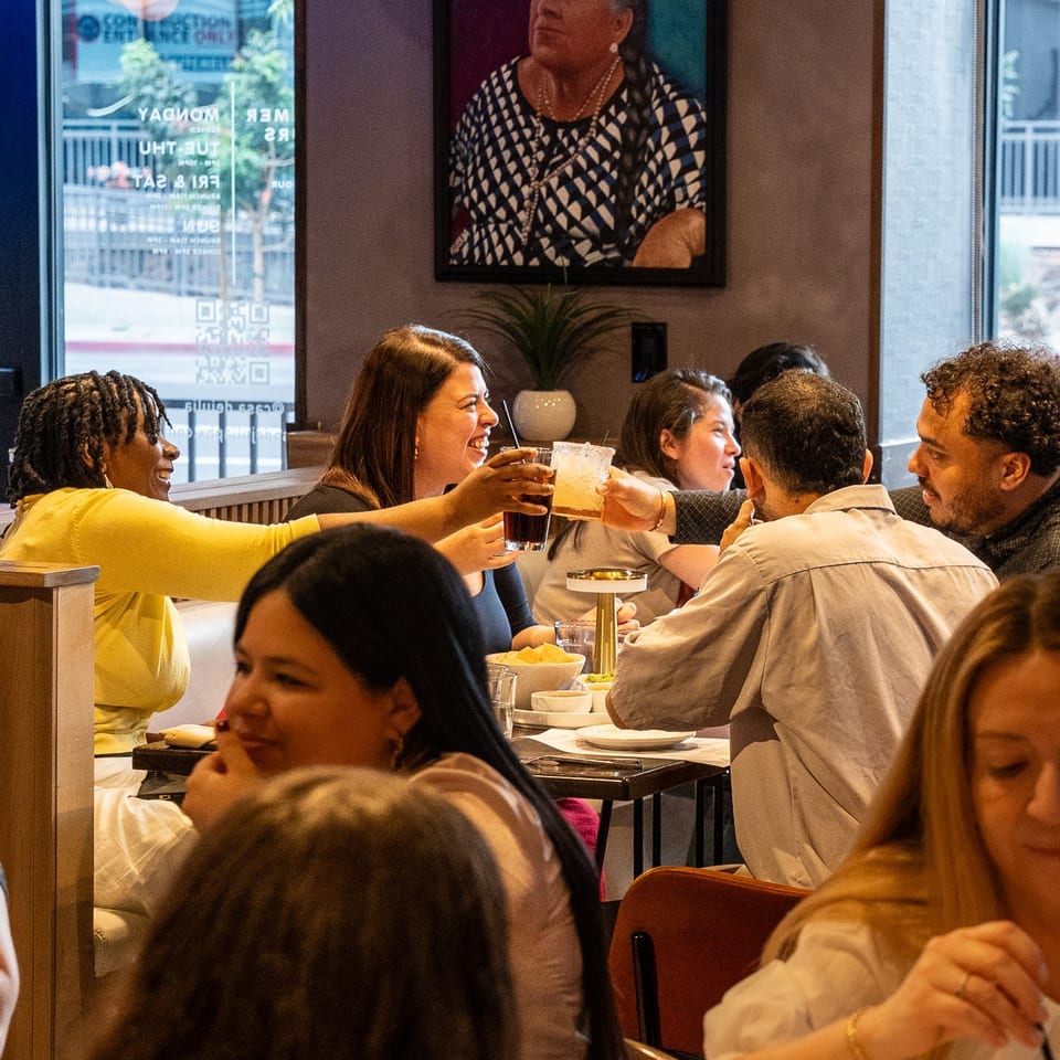 Diverse group of people dining and toasting drinks in a lively restaurant with a colorful painting.