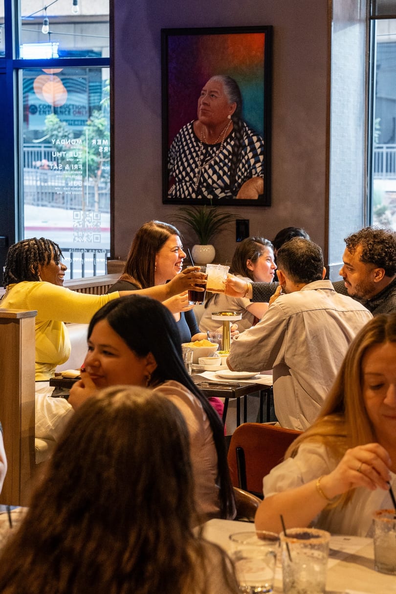 Diverse group of people dining and toasting drinks in a lively restaurant with a colorful painting.